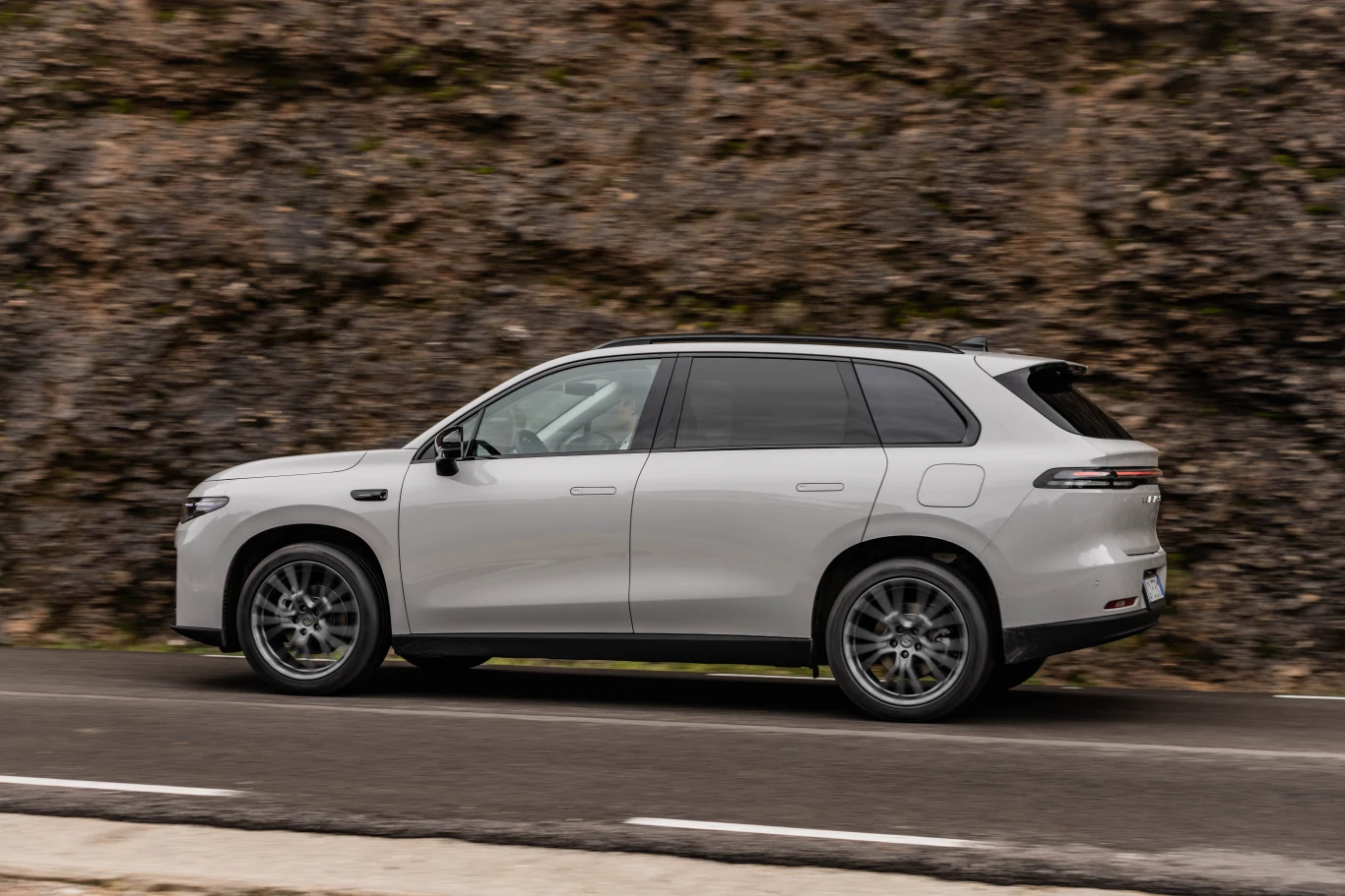 Srebrny SUV w ruchu, elegancki profil oraz nowoczesne reflektory i felgi podczas jazdy asfaltową drogą wzdłuż skalistego zbocza.