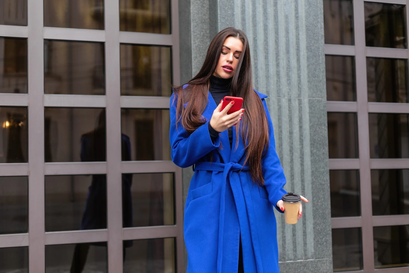 Kobieta w niebieskim płaszczu z długimi rozpuszczonymi włosami patrzy na czerwony smartfon, trzymając w drugiej ręce kubek z kawą.