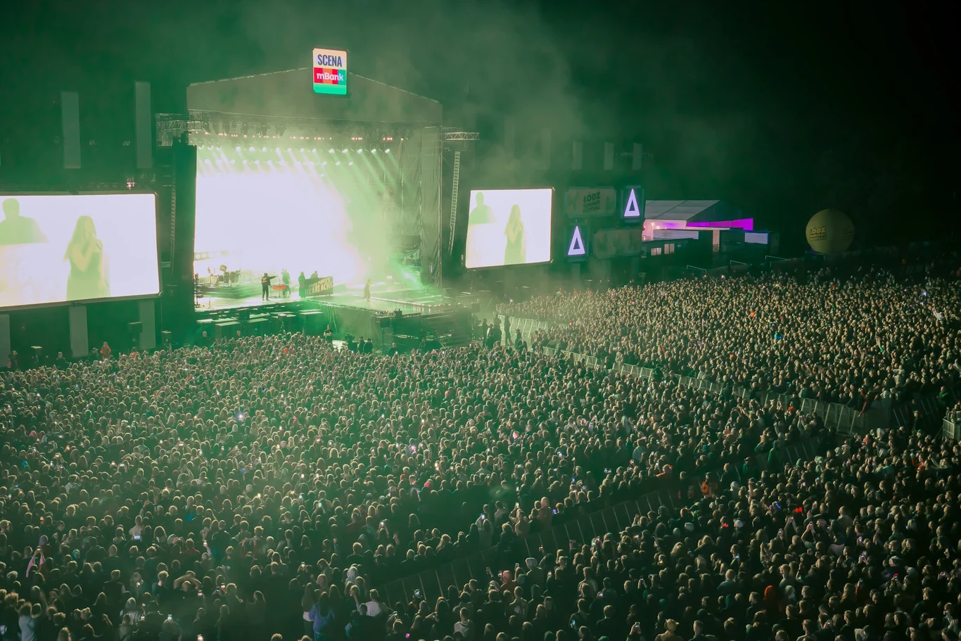 Tłum ludzi na dużym plenerowym koncercie przed jasno oświetloną sceną z wielkoformatowymi ekranami po bokach.