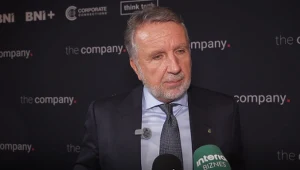 Mężczyzna w garniturze i krawacie udzielający wywiadu przed ścianką z logotypami różnych firm, trzymający mikrofony z widocznym logo „Interia Biznes”.
