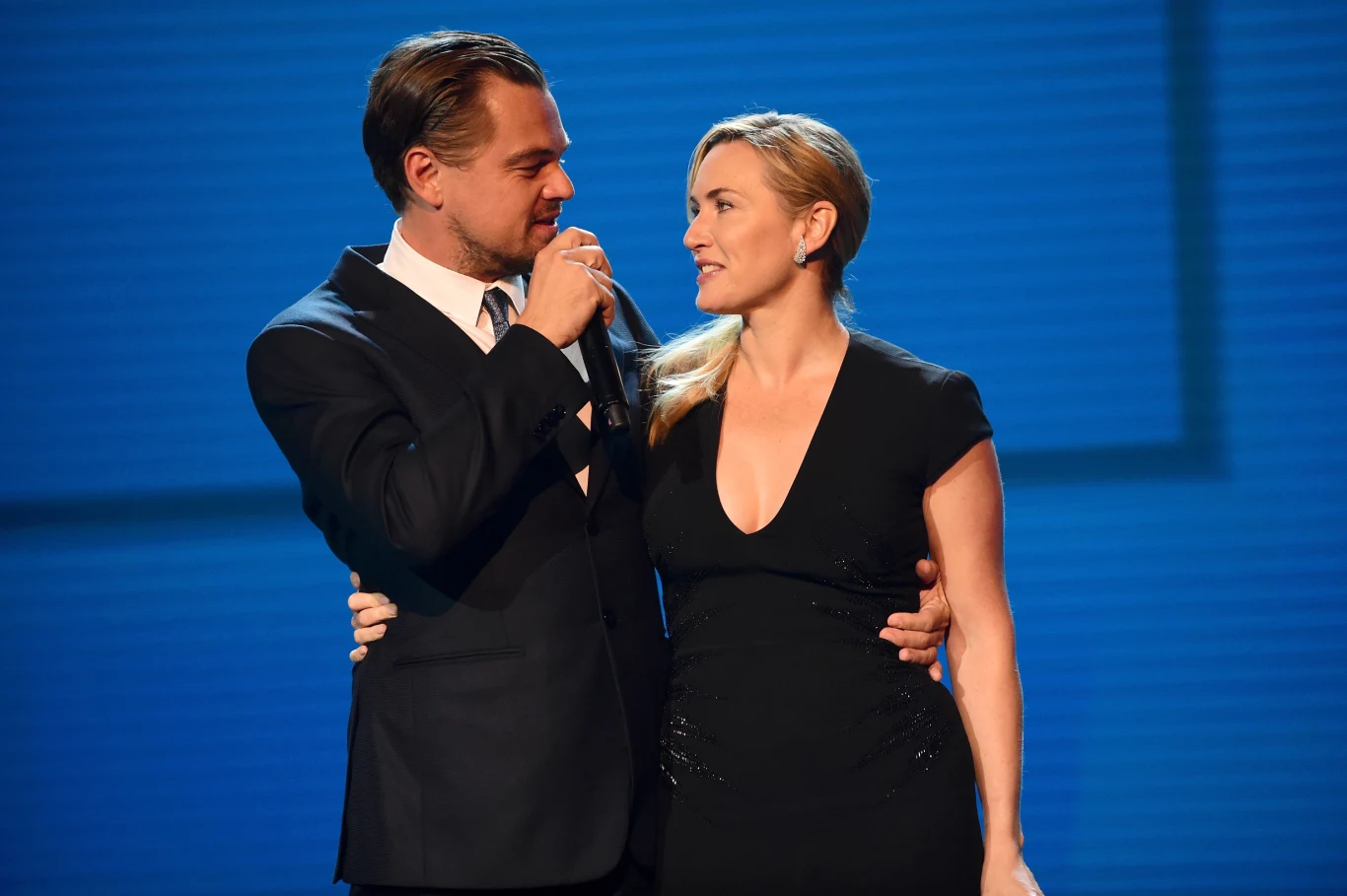 Leonardo DiCaprio i Kate Winslet Dwoje elegancko ubranych ludzi w wieczorowych strojach obejmujących się na scenie, mężczyzna trzyma mikrofon i patrzy z uśmiechem na kobietę.
