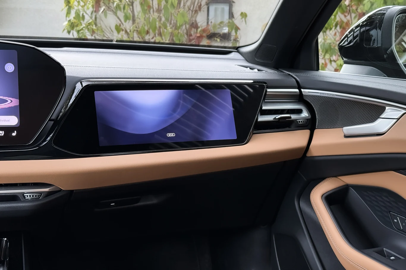 Kokpit samochodu marki Audi z dużym ekranem multimedialnym na desce rozdzielczej, jasnobrązowe wykończenia wnętrza oraz eleganckie, nowoczesne panele drzwiowe.