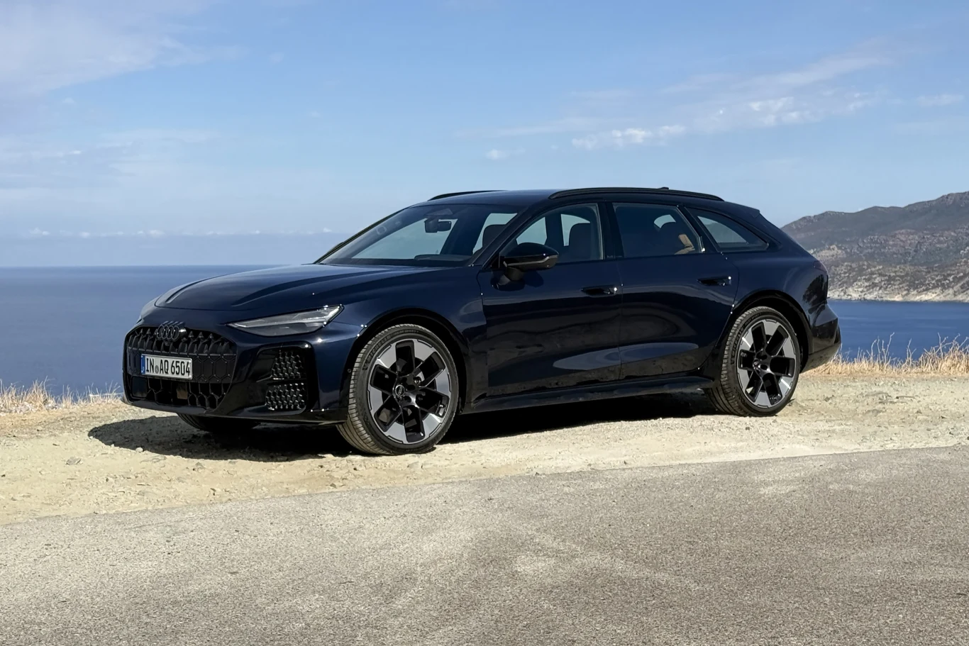 Czarne kombi Audi zaparkowane przy klifie z widokiem na morze i górzyste wybrzeże w tle.