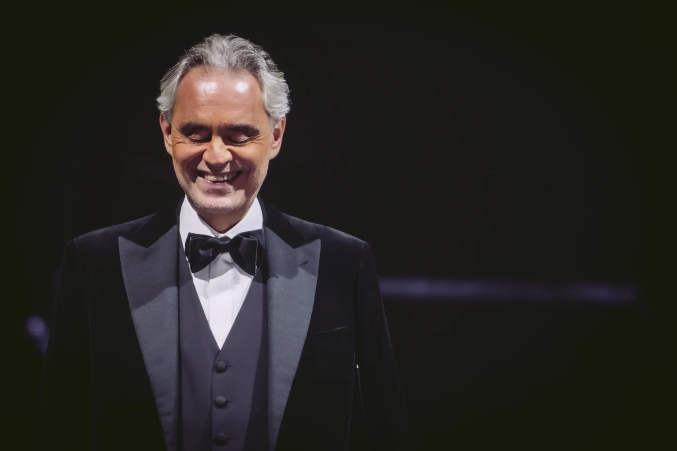 Andrea Bocelli ma w Polsce wielu fanów Andrea Bocelli w eleganckim smokingu, biała koszula i mucha, na ciemnym tle.