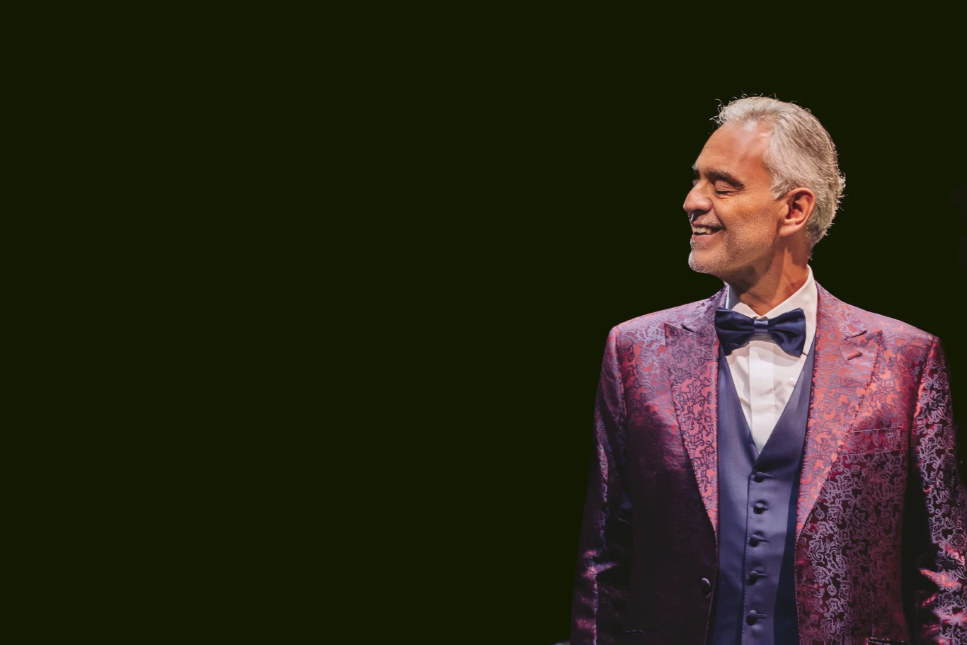 Andrea Bocelli przyjedzie do Chorzowa w ramach trasy z okazji 30-lecia płyty "Romanza" Elegancki mężczyzna w bordowej, błyszczącej marynarce i granatowej muszce, patrzący z uśmiechem w bok na ciemnym tle.