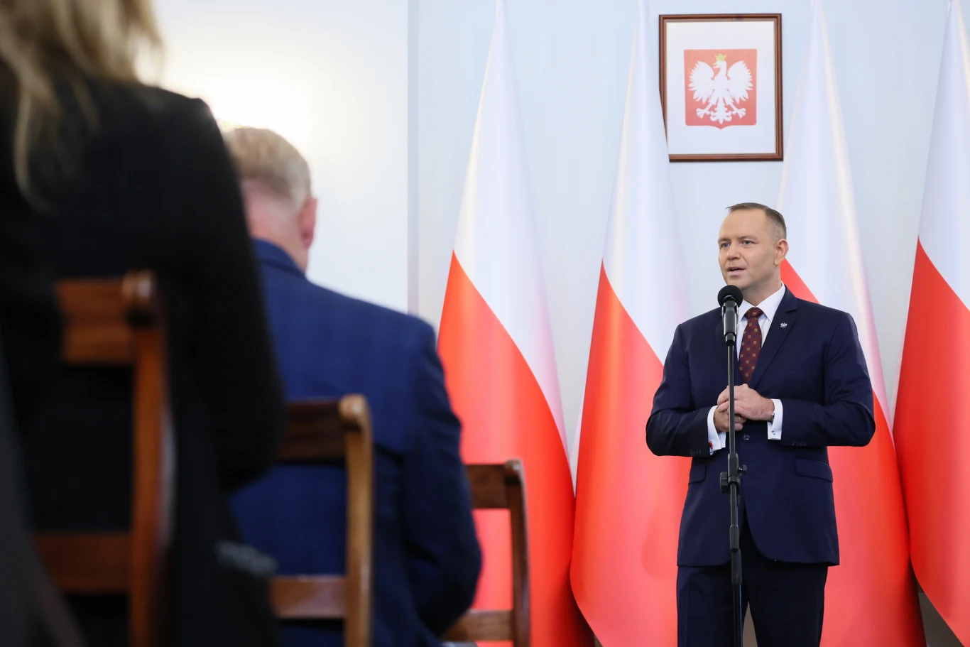 Prezydent Karol Nawrocki Prezydent Karol Nawrocki przemawia przy mikrofonie, w tle polskie flagi i godło, przed nim dwie osoby.