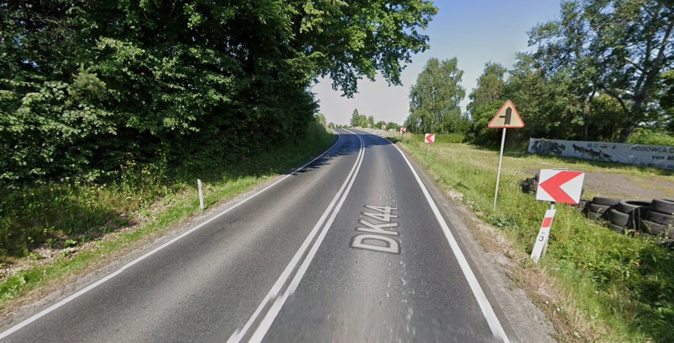 Dk. 44 m. Jaśkowice. GDDKiA planuje dużą modernizację. Jednopasmowa DK44 w Jaśkowicach otoczona zielenią, łagodny prawy zakręt, znaki ostrzegawcze i barierki.