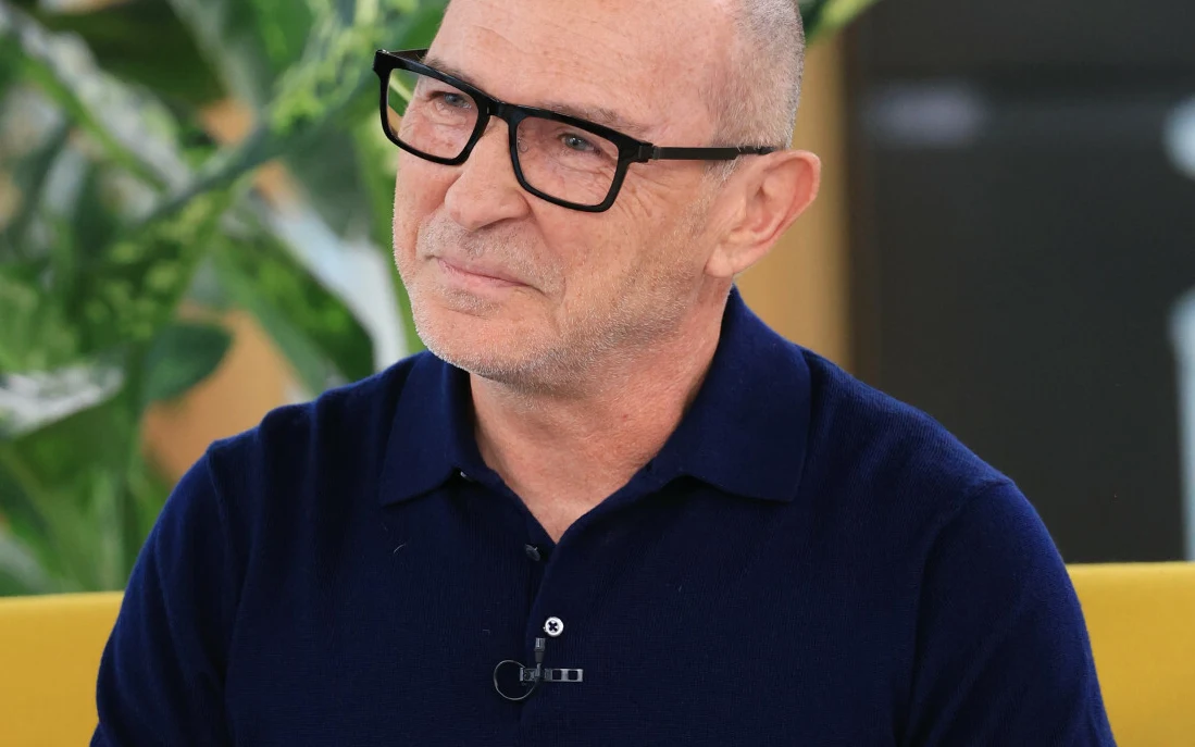 Andrzej Krzywy Andrzej Krzywy w granatowej koszulce polo i okularach, siedzący na żółtej sofie na tle roślin.