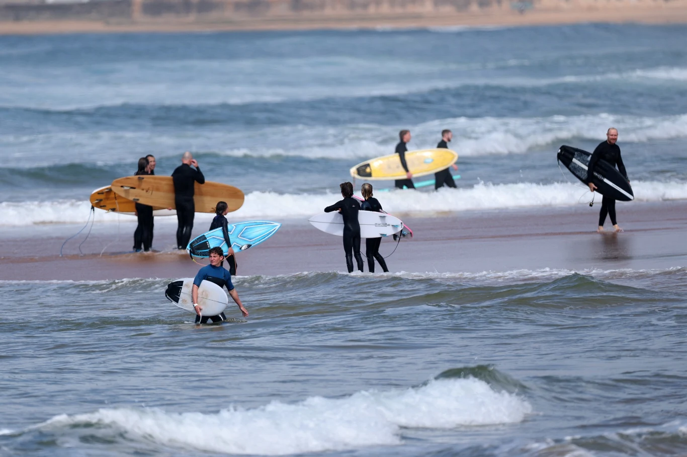 Grupa surferów z deskami przygotowuje się do wejścia do wody, część osób już stoi w płytkiej części morza, a w tle widoczne są niewielkie fale.