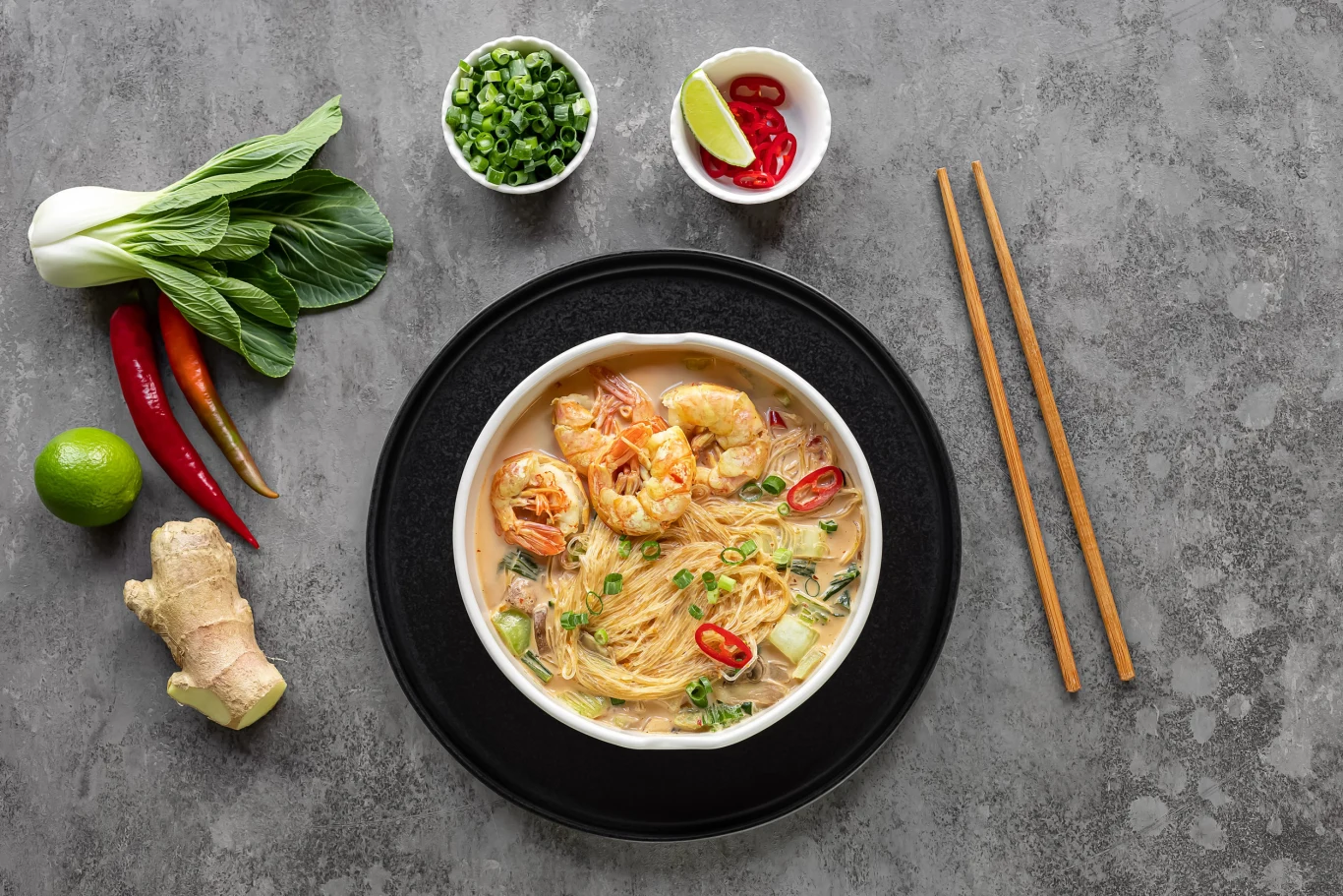 Zupa laksa to jedna z lepszych zup azjatyckich Miska zupy laksa z makaronem ryżowym, krewetkami, papryczkami chili i warzywami, pałeczki oraz składniki obok.