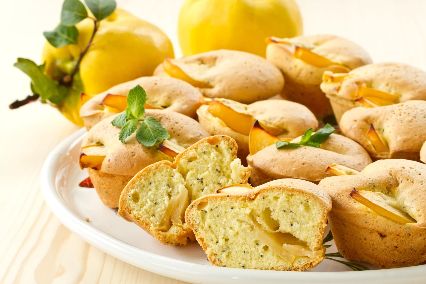 Bałkańskie babeczki z pigwą i makiem. Muffinki z pigwą i makiem na białym talerzu, udekorowane listkami mięty, w tle widoczne owoce pigwy.