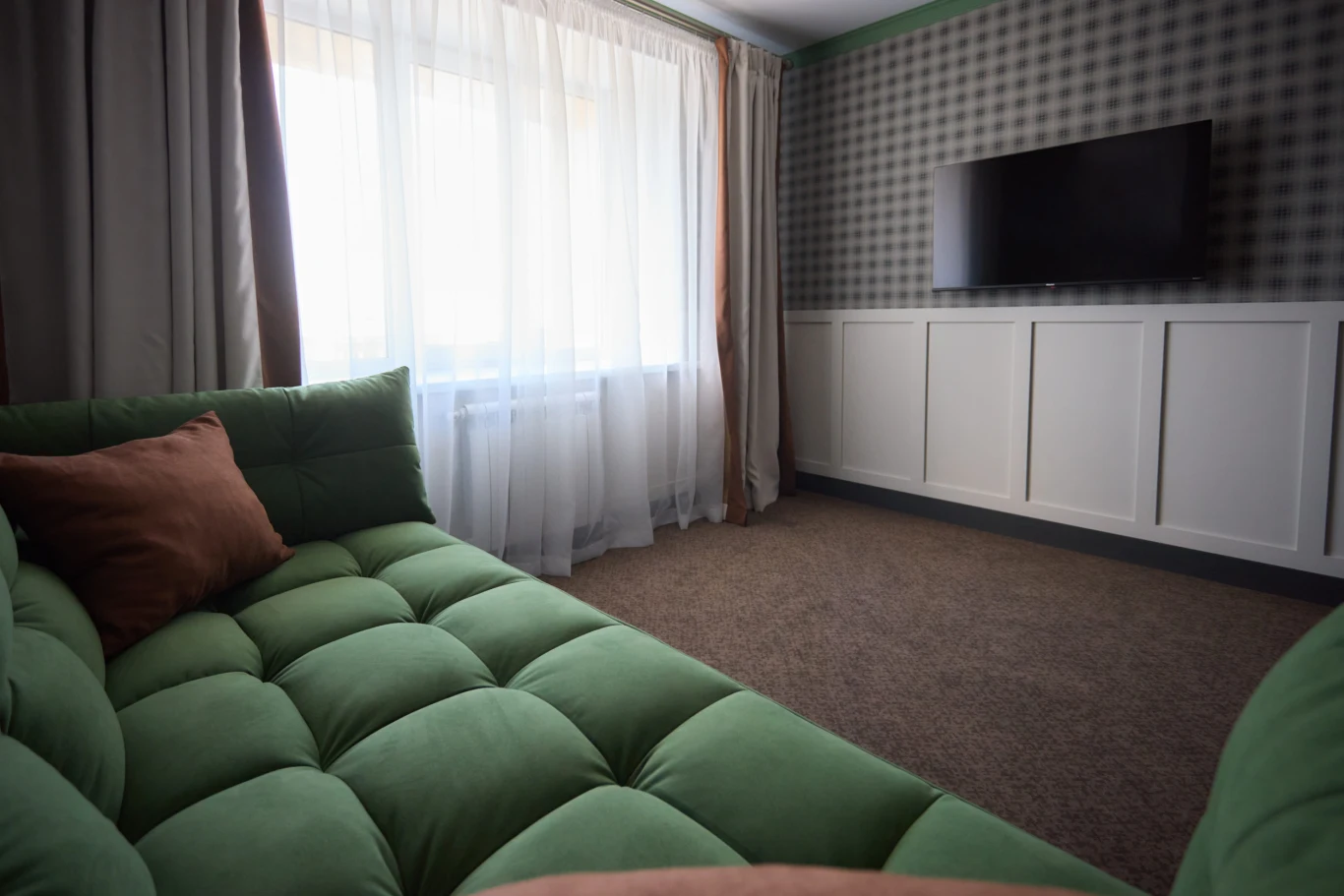 Zielona, pikowana sofa z brązową poduszką stoi naprzeciwko ściany z telewizorem zawieszonym na geometrycznej tapecie i jasną lamperią, a duże okno z białymi firanami wpuszcza do wnętrza naturalne światło.