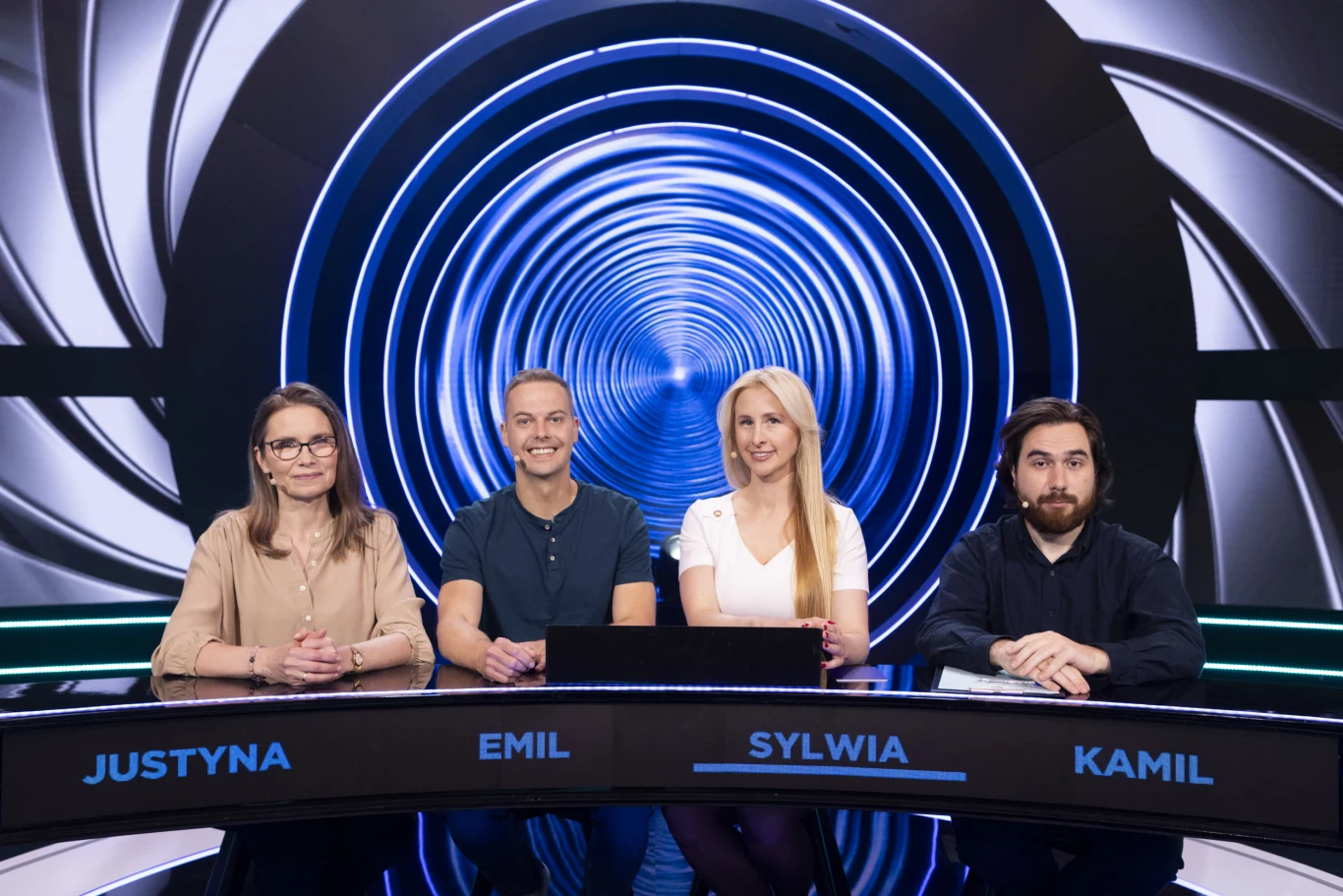 Cztery osoby siedzą przy okrągłym stole w studiu telewizyjnym na tle niebieskiego tunelu świetlnego, każda z nich ma przed sobą tabliczkę z imieniem.