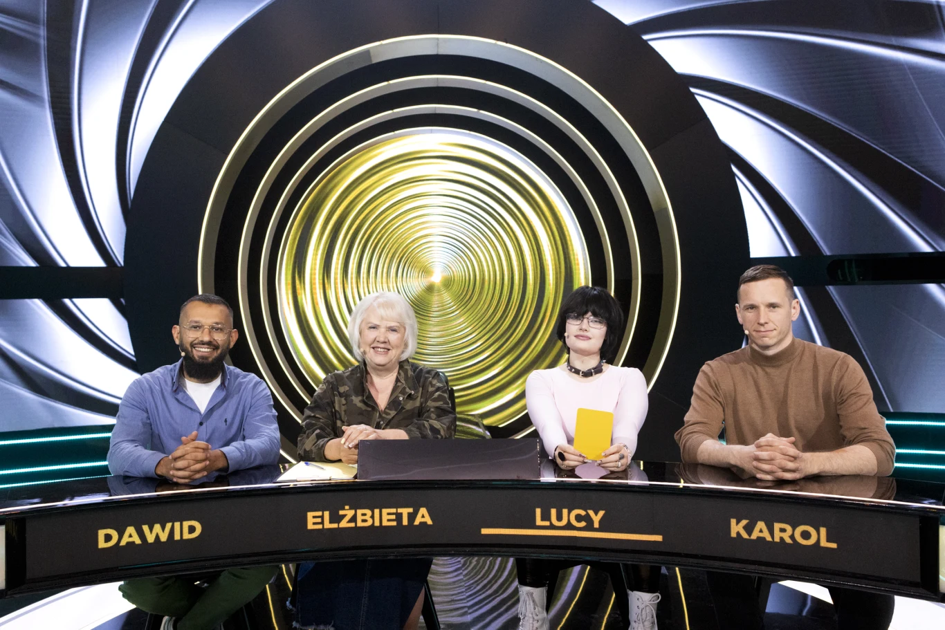 Czworo osób siedzi za nowoczesnym, półokrągłym stołem w studiu telewizyjnym z futurystyczną, spiralną dekoracją w tle, a przed każdym widnieje tabliczka z imieniem.