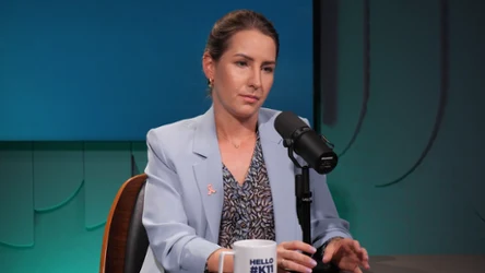 Kobieta w jasnoniebieskiej marynarce siedzi przy stole podczas nagrania podcastu, trzyma długopis i patrzy poważnie na rozmówcę, przed nią stoi mikrofon oraz biały kubek z napisem.
