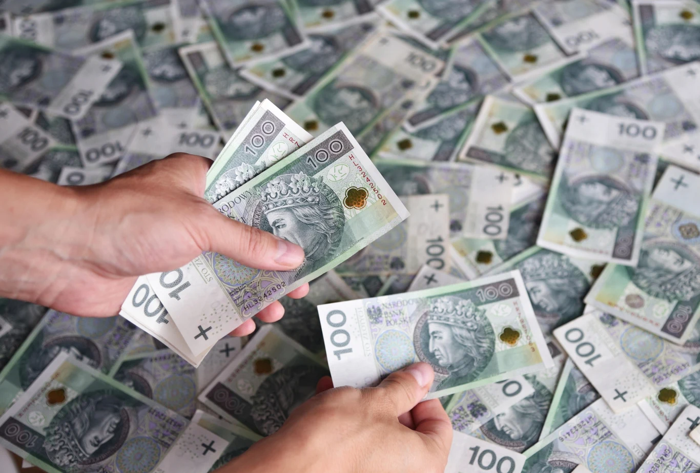 Są pieniądze na pomysły mieszkańców Alwerni. Gmina przygotowała 100 tys. zł. Dwie dłonie trzymają i liczą banknoty 100 zł na tle rozłożonych banknotów, kontekst budżetu obywatelskiego.