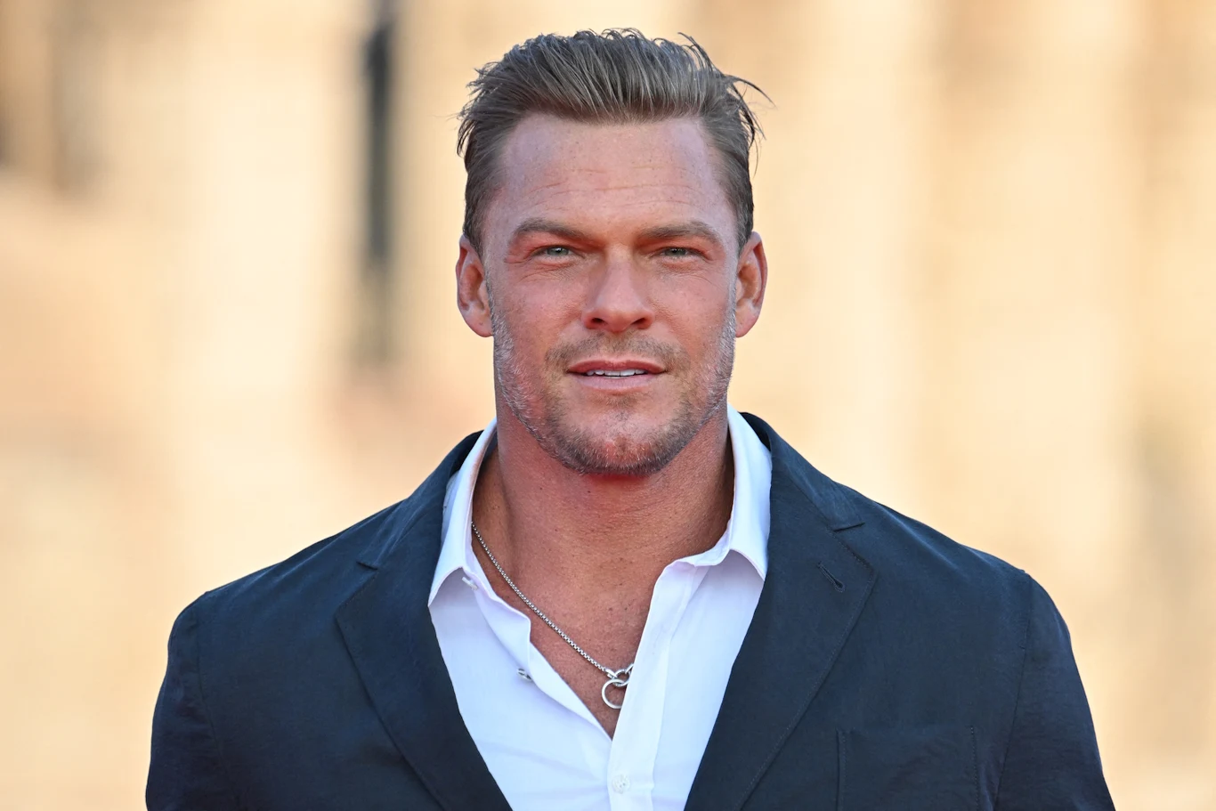 Alan Ritchson Alan Ritchson w granatowej marynarce i białej koszuli, krótkie włosy, srebrny łańcuszek na szyi.