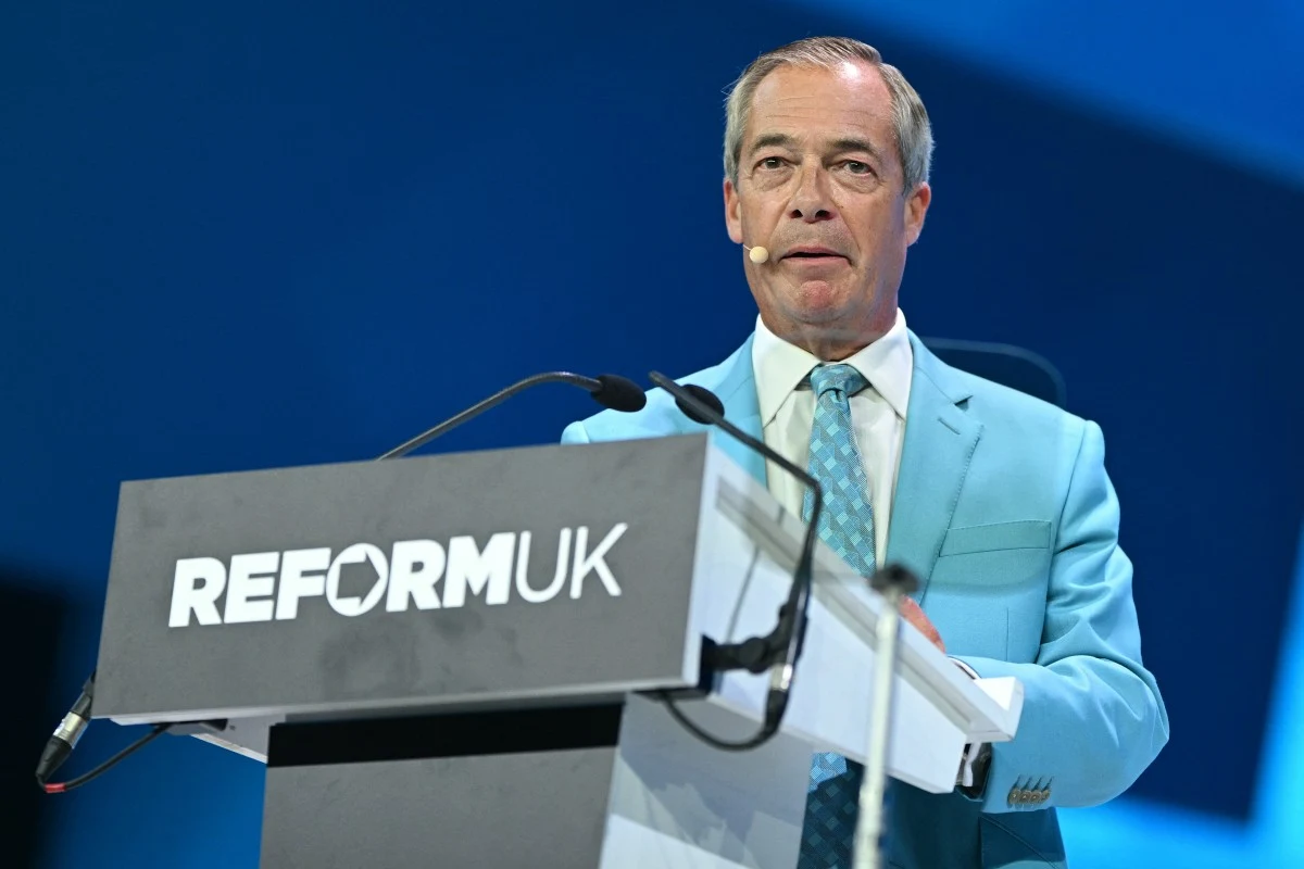 Lider partii Reform UK Nigel Farage Nigel Farage w jasnoniebieskim garniturze przemawia przy mównicy z logo Reform UK na tle niebieskiej scenografii