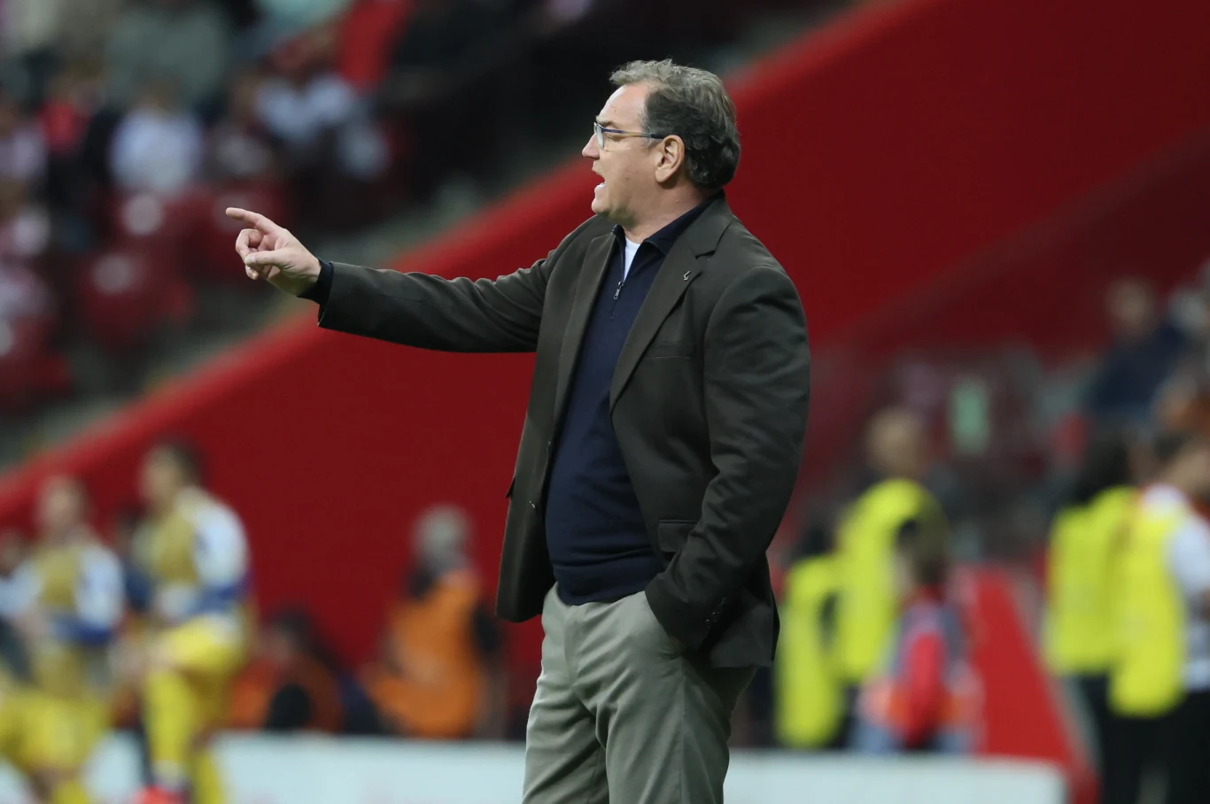 Mężczyzna w garniturze gestykuluje, stojąc na tle czerwonych trybun stadionu, wokół niego widoczni są zawodnicy lub członkowie sztabu sportowego w żółtych kamizelkach.
