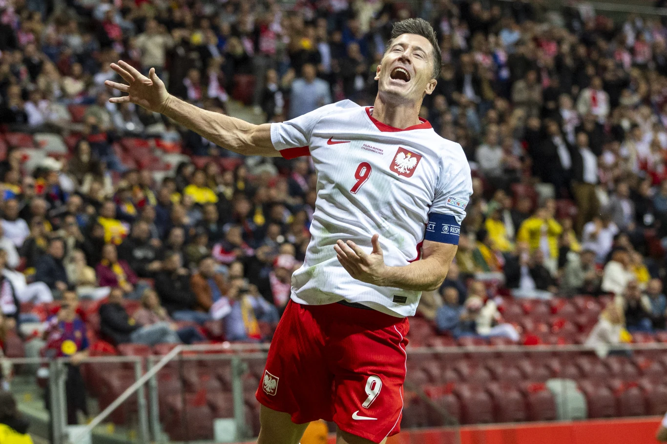 Robert Lewandowski Piłkarz w białej koszulce z numerem 9 i opaską kapitańską świętuje zdobycie bramki na stadionie podczas meczu reprezentacji Polski, z trybunami wypełnionymi kibicami w tle.