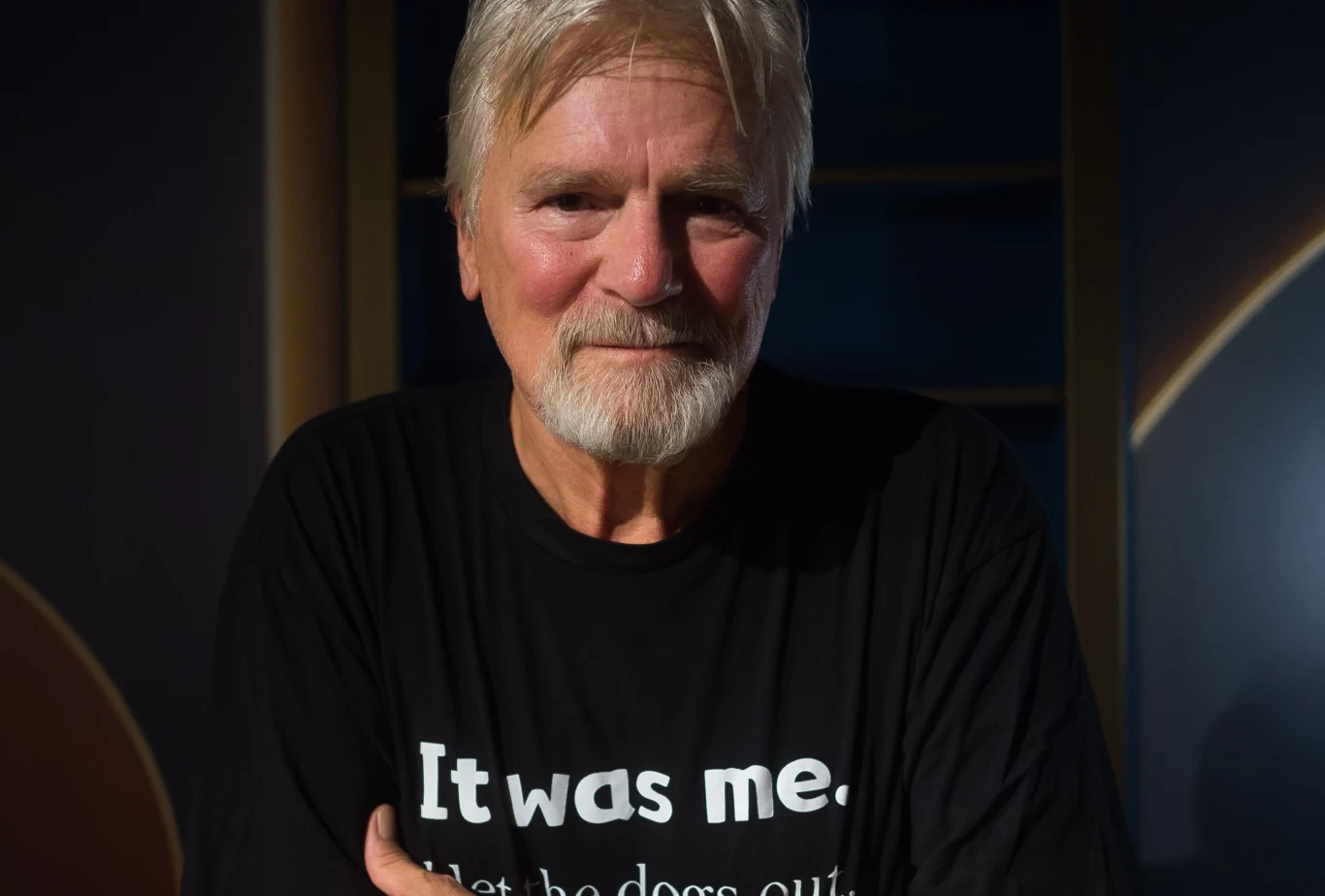 Starszy mężczyzna z siwymi włosami i brodą, ubrany w czarną koszulkę z napisem 'It was me.'