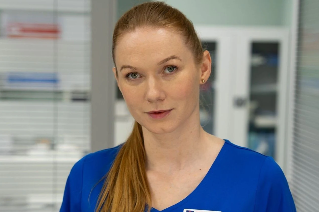Wiktoria Manuela Consalida (Katarzyna Dąbrowska) Kobieta o rudych włosach spiętych w kucyk, ubrana w niebieski uniform medyczny, stoi w jasnym pomieszczeniu