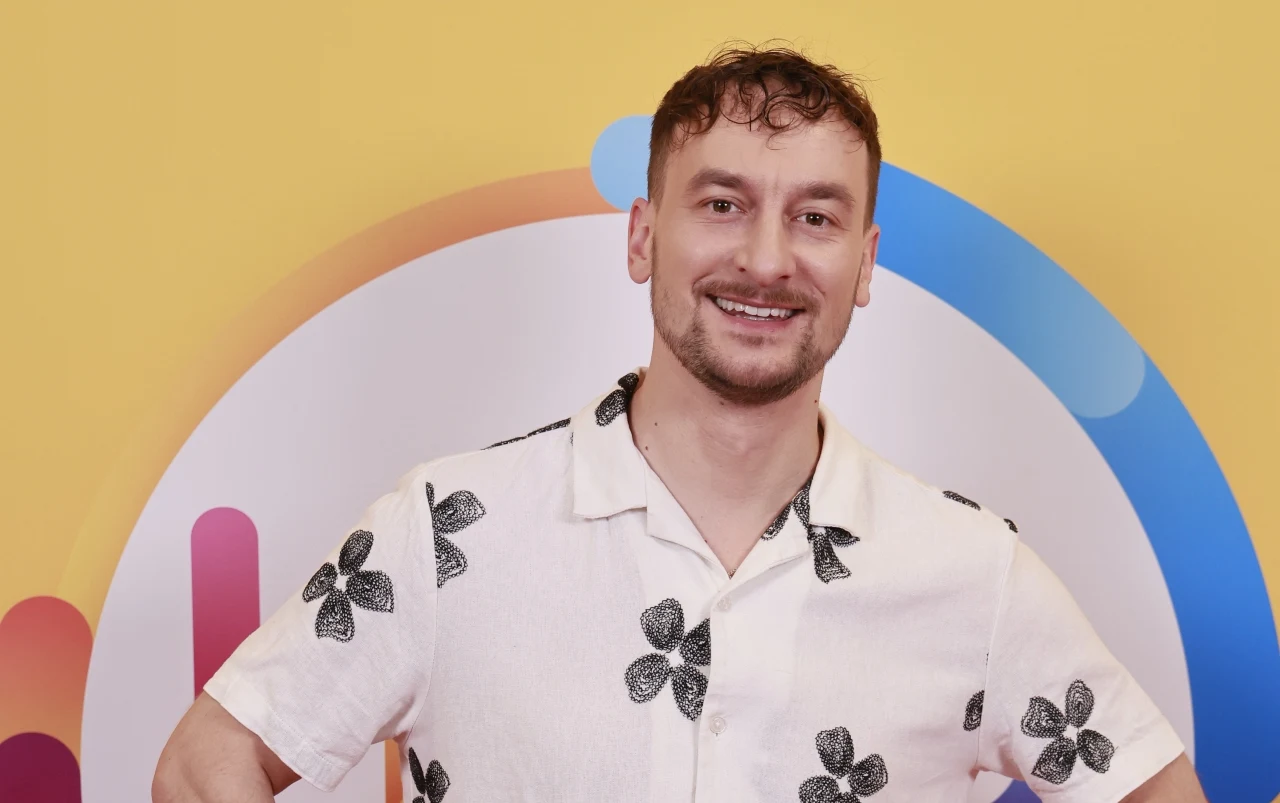 Przemysław Niedzielski, czyli przehehemek o tym, skąd bierze pomysły na rozśmieszanie ludzi Przemysław Niedzielski w koszuli w czarne kwiaty na tle kolorowej grafiki z okrągłymi kształtami.