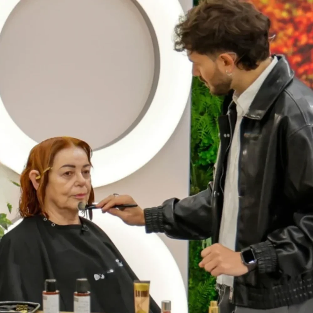 Kobieta z widocznymi sztucznymi uszami siedzi na fotelu fryzjerskim, a mężczyzna w czarnej kurtce wykonuje makijaż pędzlem na jej twarzy.