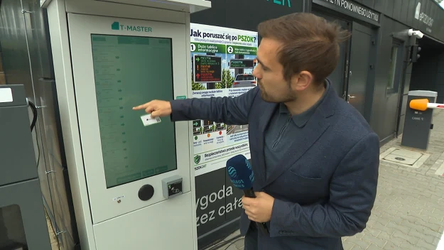 Mężczyzna w garniturze obsługujący duży interaktywny panel dotykowy z kartą w ręce, trzyma mikrofon i wskazuje palcem na ekran automatu umieszczonego na chodniku przy budynku.