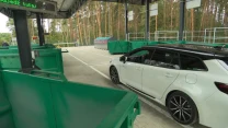 Białe kombi zaparkowane obok dużych zielonych pojemników na odpady na terenie punktu selektywnej zbiórki odpadów, w tle drzewa i ogrodzenie.