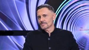 Mężczyzna w ciemnej marynarce siedzi na tle futurystycznych, okrągłych wzorów świetlnych w odcieniach fioletu i błękitu.