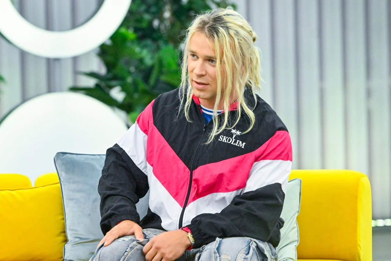 Skolim w "halo tu polsat" Młody mężczyzna z blond dredami ubrany w sportową kurtkę w biało-czarno-różowe pasy siedzi na żółtej sofie, zrelaksowany oparty o poduszkę.