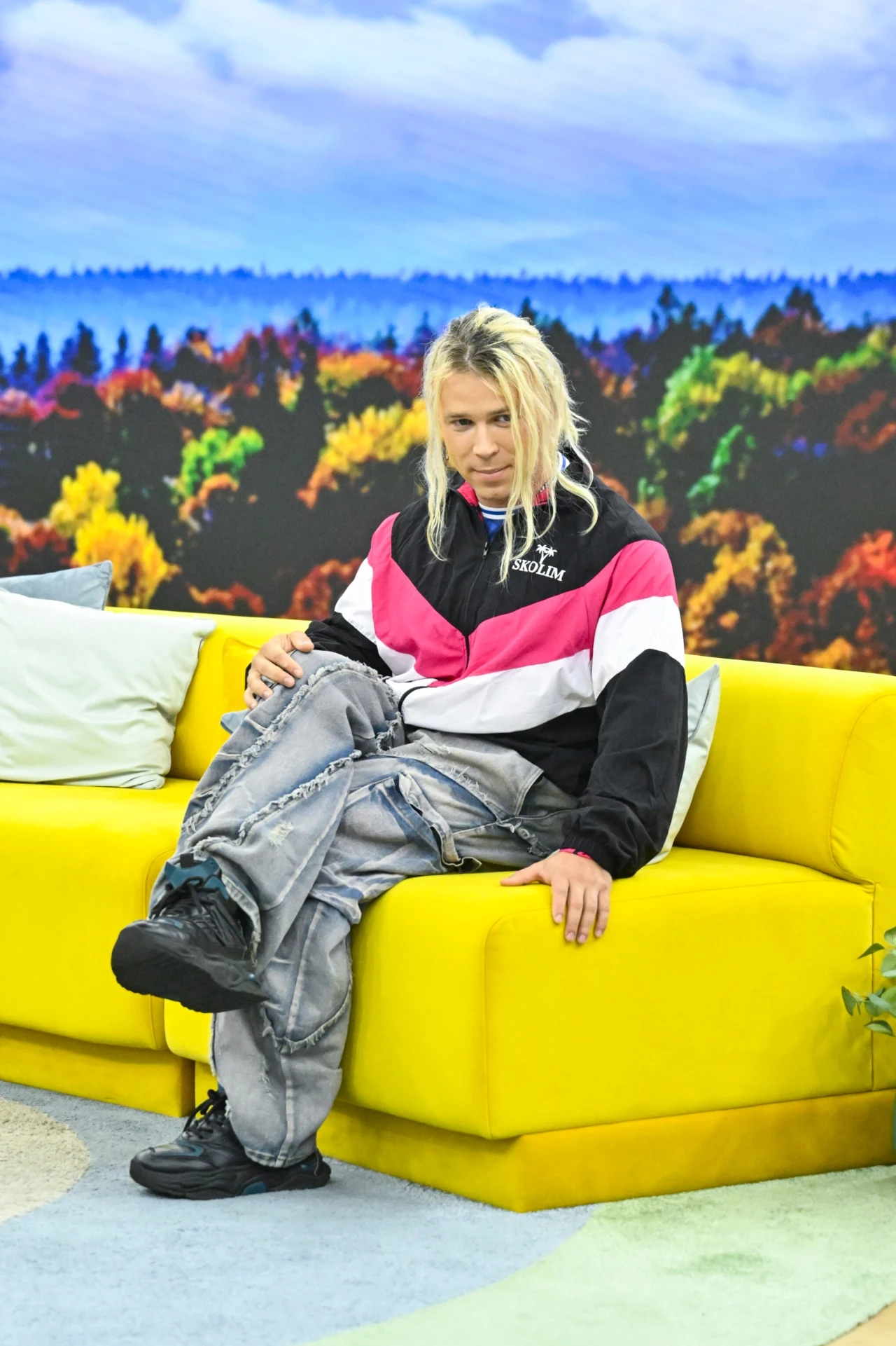 Skolim w "halo tu polsat" Młoda osoba z blond dreadami siedzi na żółtej sofie, ubrana w sportową kurtkę w kolorach czarnym, różowym i białym oraz szerokie szare spodnie, na tle kolorowej jesiennej panoramy w studiu telewizyjnym.