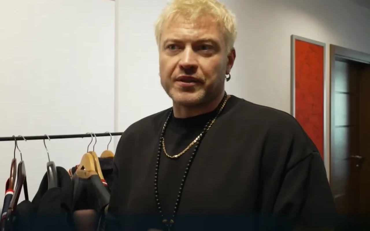 Łozo pokazuje swoją garderobę w "halo tu polsat" Mężczyzna z blond włosami ubrany w czarną bluzę i naszyjniki, stojący przy stojaku z ubraniami w jasnym pomieszczeniu.