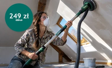 Kobieta w okularach ochronnych i maseczce szlifuje gładź na skośnym suficie poddasza szlifierką na teleskopie