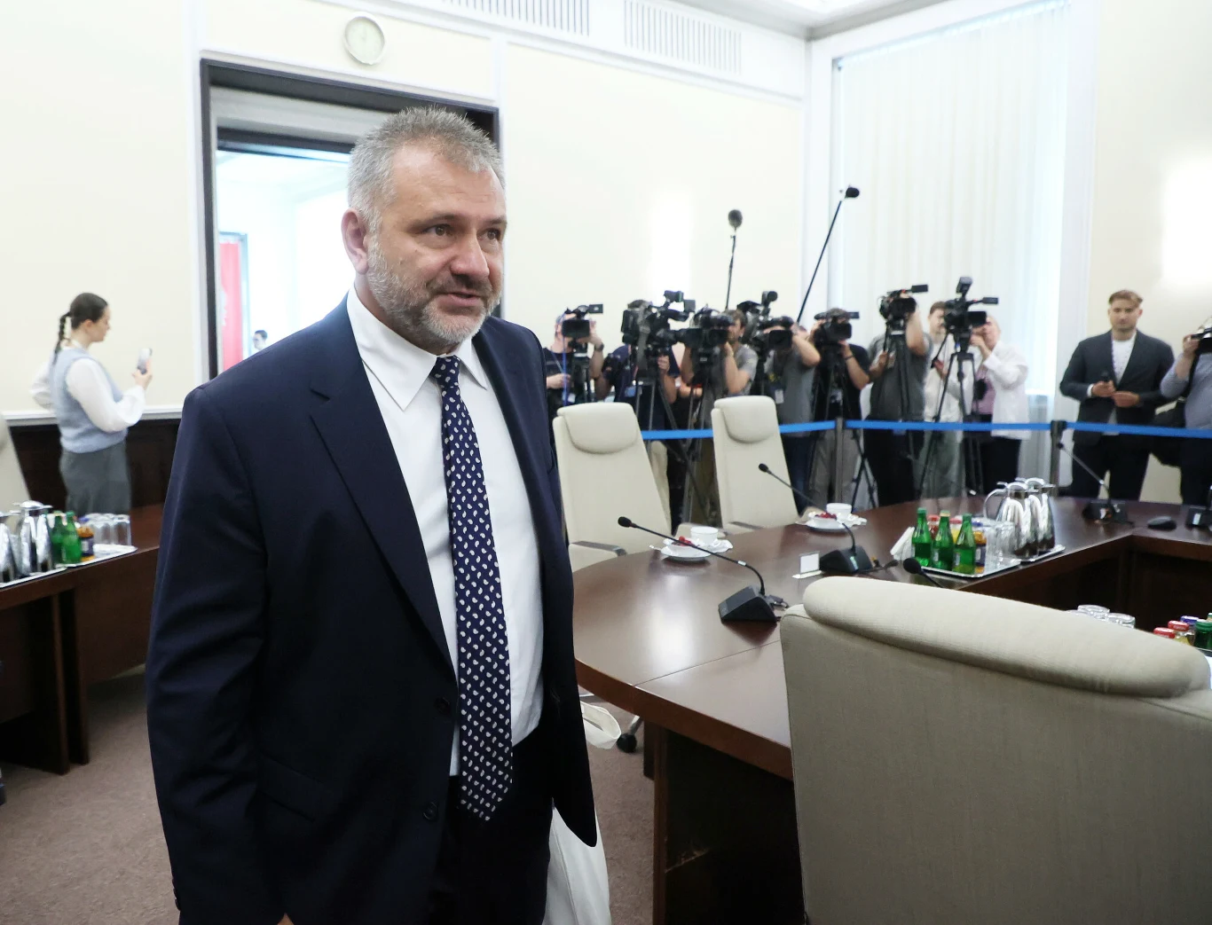 Mężczyzna w garniturze stoi przy konferencyjnym stole, wokół którego ustawiono kilka foteli, a w tle grupa fotoreporterów z aparatami i kamerami dokumentuje wydarzenie.