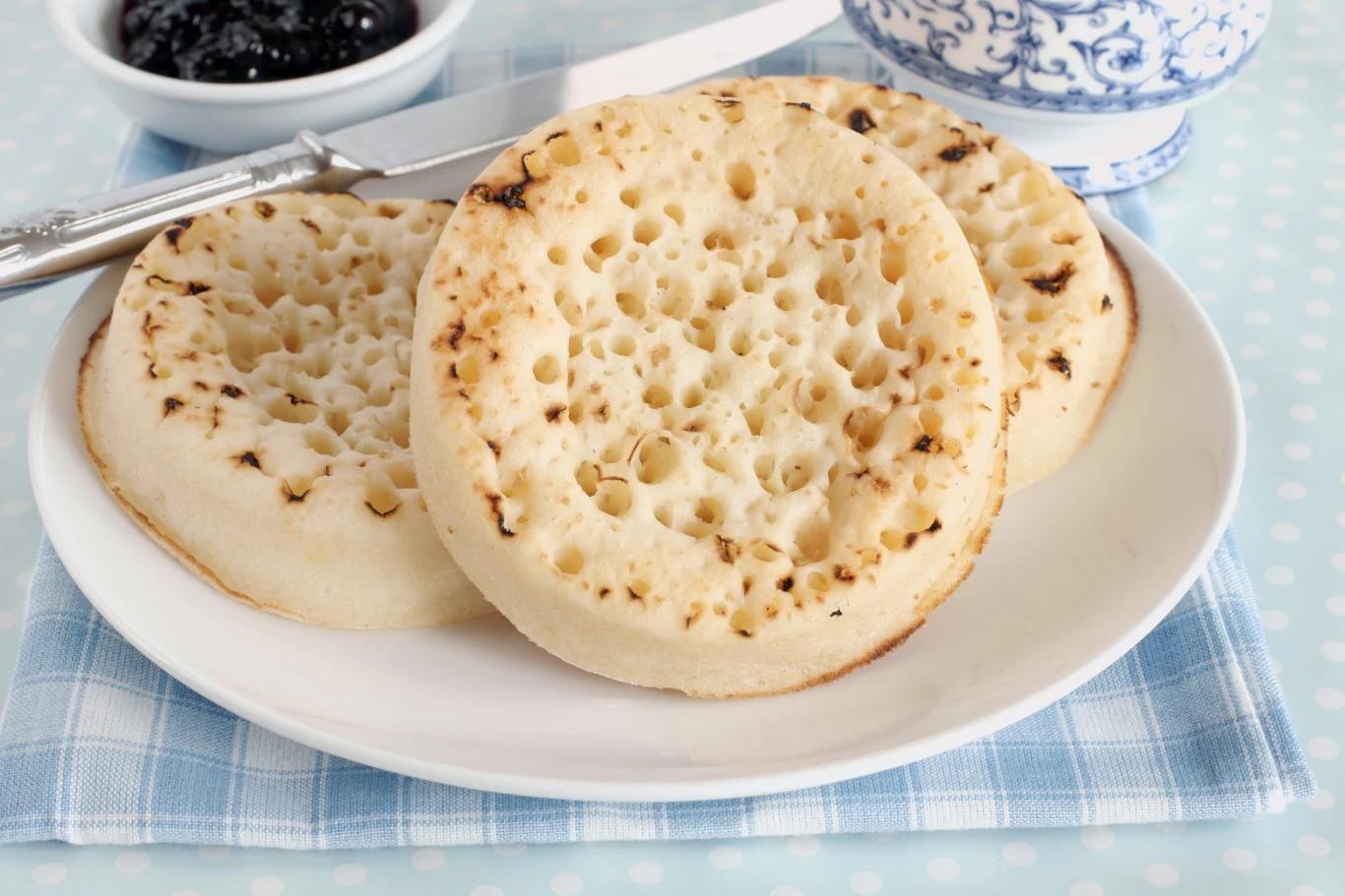 Charakterystyczne dziurki na crumpets doskonale wchłaniają masło Crumpets z dziurkami na białym talerzu, obok miseczka z dżemem.