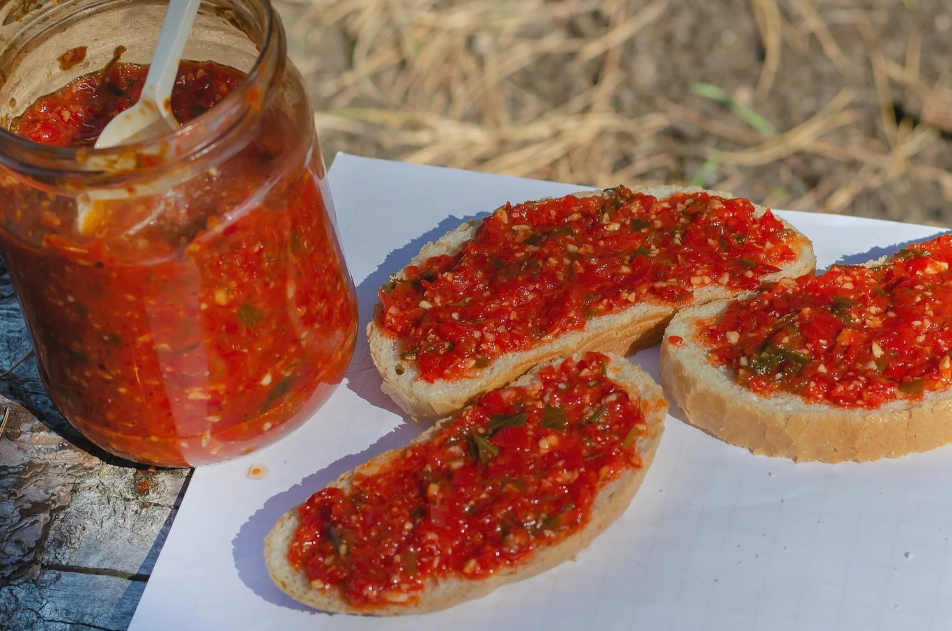 Gęsta i aromatyczna pasta z pieczonej papryki. na kanapki nie ma nic pyszniejszego Trzy kromki chleba posmarowane pastą z pieczonej papryki, obok słoik pasty z łyżką.