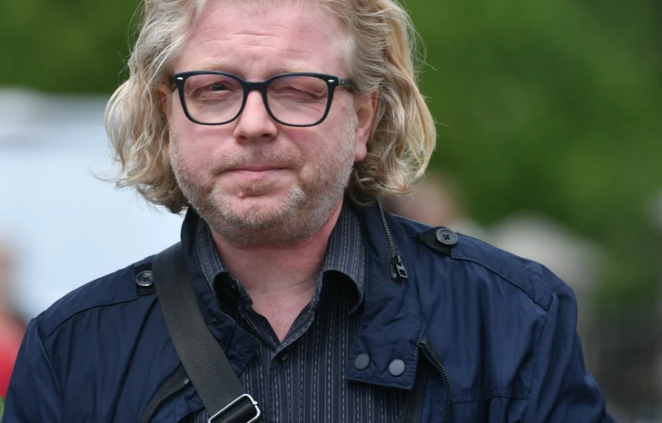 Przemysław Branny Mężczyzna z długimi blond włosami, noszący okulary i ciemną kurtkę, patrzący wprost przed siebie, z jednym okiem przymrużonym.
