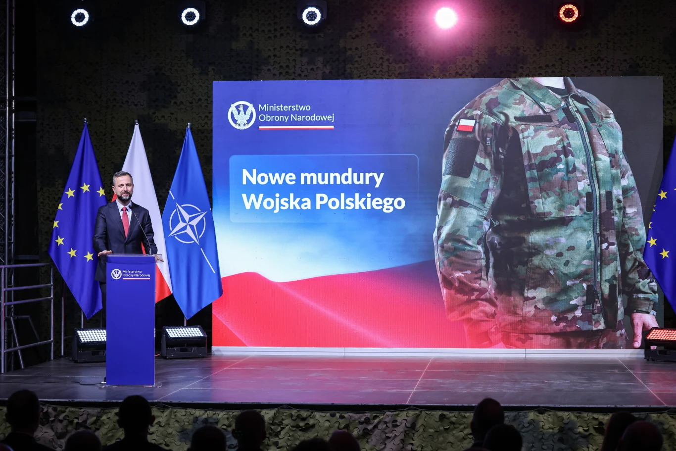 Minister obrony narodowej prezentuje nowe mundury Wojska Polskiego podczas oficjalnej konferencji, na scenie widoczne są flagi Unii Europejskiej, NATO i Polski oraz duży ekran z grafiką żołnierza w nowym mundurze.