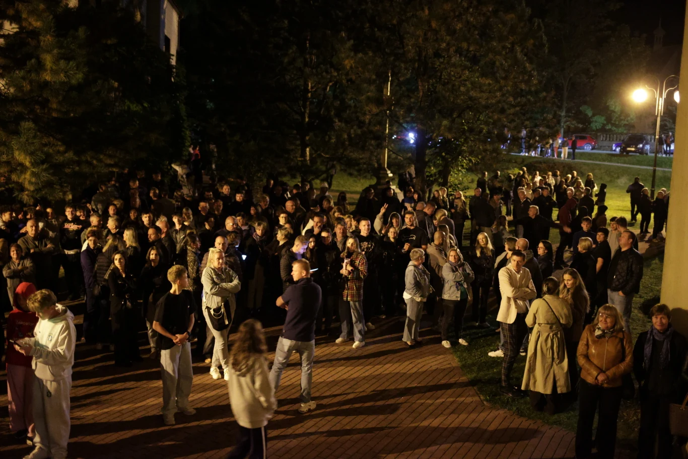 Protest przed kościołem pw. Świętego Wawrzyńca w Grojcu Duża grupa ludzi zgromadzona na zewnątrz po zmroku, większość stoi na chodniku i trawniku w pobliżu lamp ulicznych oraz drzew, część osób rozmawia lub robi zdjęcia.