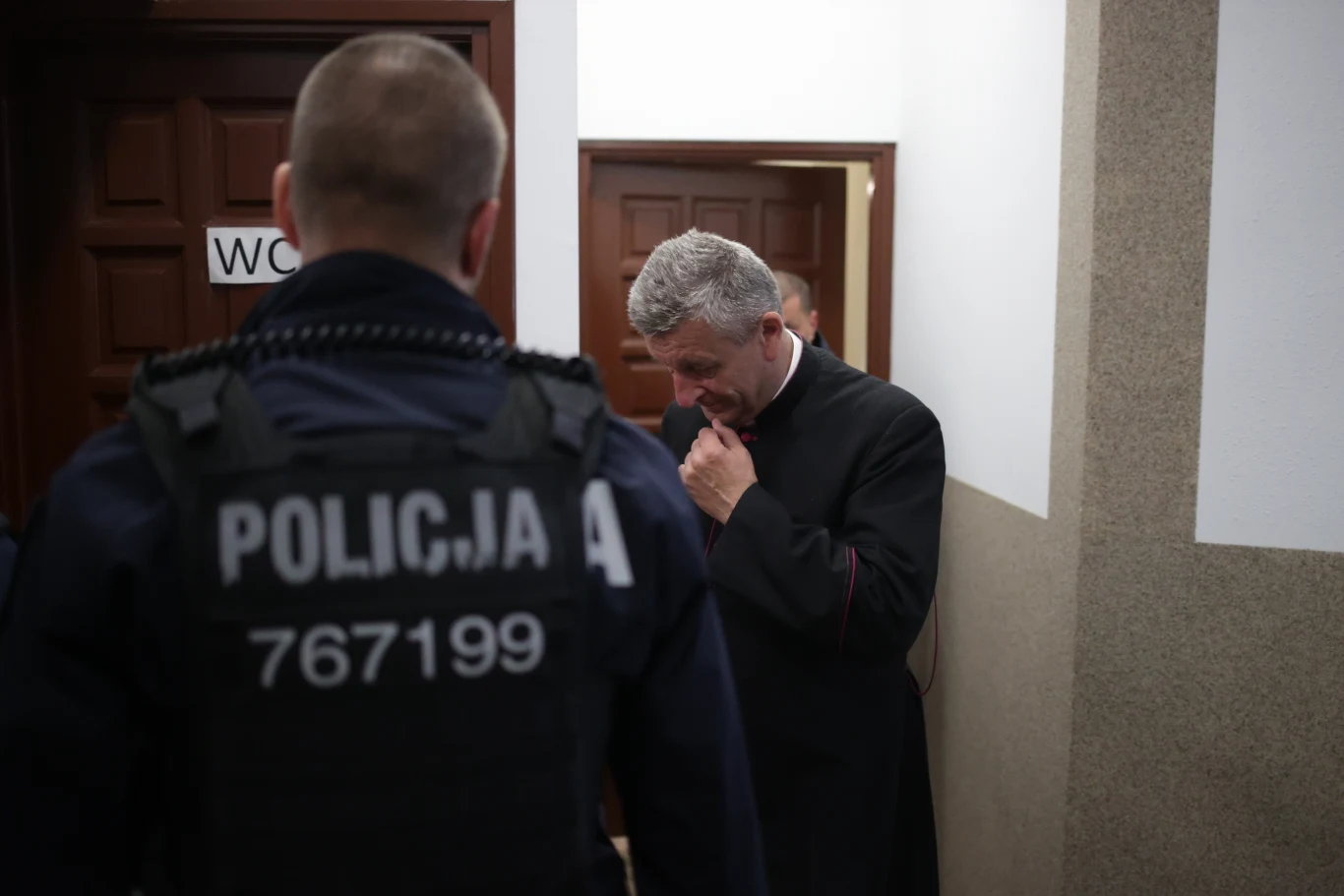 Parafianie protestowali. Biskup nie mógł wyjść z kościoła Policjant stojący tyłem w kamizelce z widocznym napisem oraz mężczyzna w sutannie, który wygląda na zaniepokojonego, znajdują się na korytarzu przed drzwiami.