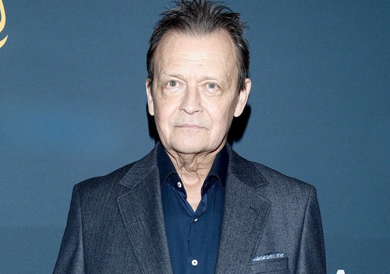 Jan Frycz Aktor Jan Frycz w granatowej koszuli i szarej eleganckiej marynarce, stoi na tle niebieskiej ścianki.