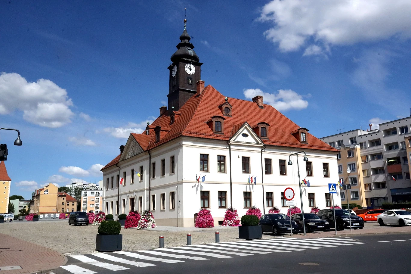 Lubin szykuje się na zmiany klimatyczne. Urzędnicy zapytali mieszkańców o zdanie. Zabytkowy ratusz w Lubinie z czerwoną dachówką i wieżą zegarową, otoczony kwiatami i zaparkowanymi autami.