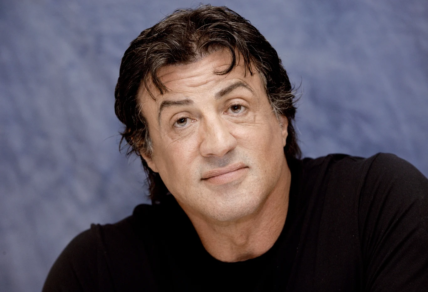 Sylvester Stallone Mężczyzna z ciemnymi, lekko falującymi włosami, ubrany w czarną koszulkę, patrzy spokojnym wzrokiem w stronę obiektywu na tle niebieskiej ściany.