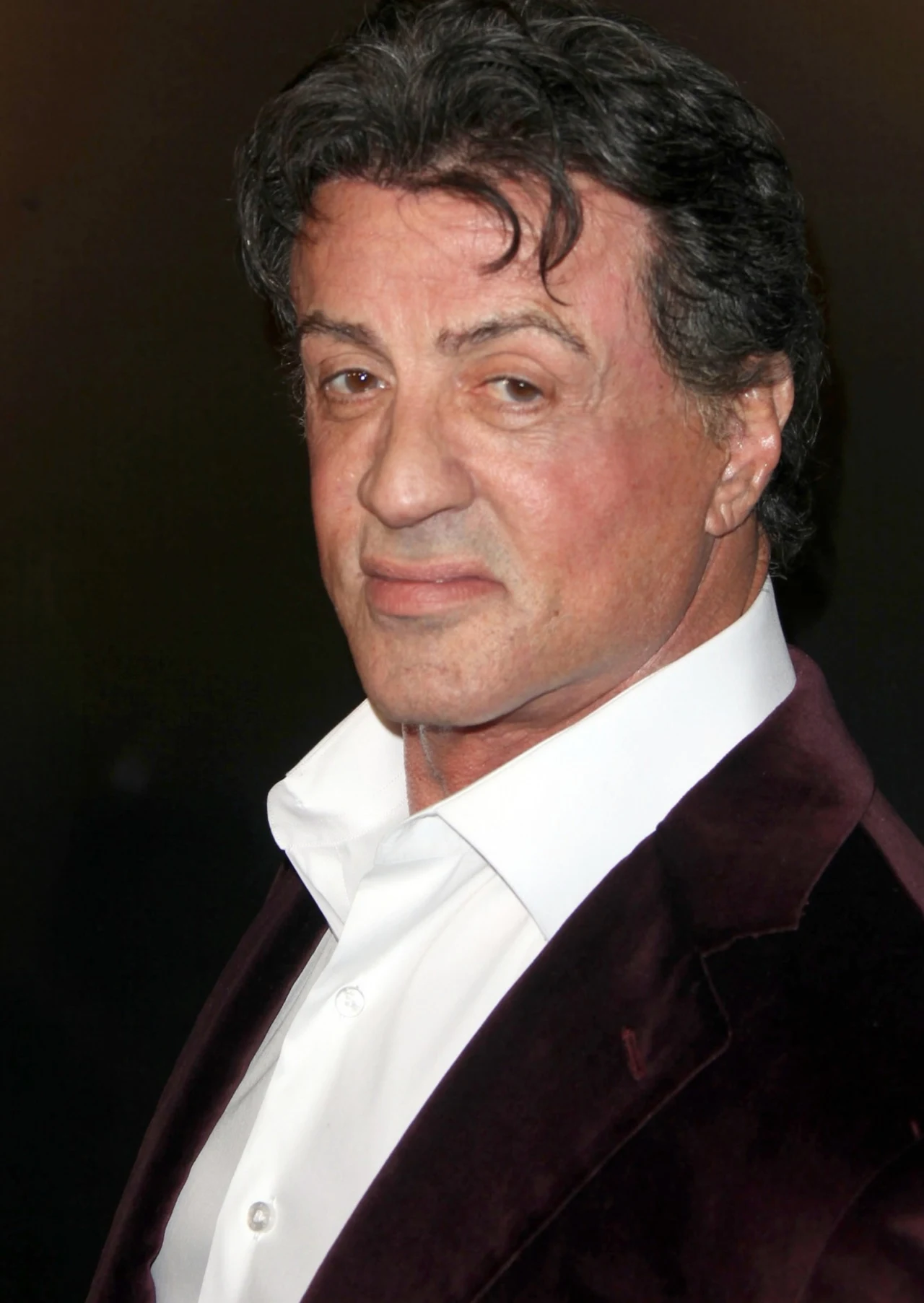 Sylvester Stallone Mężczyzna w średnim wieku z ciemnymi, falującymi włosami, ubrany w ciemną marynarkę i białą koszulę, patrzy lekko w bok z delikatnym uśmiechem.