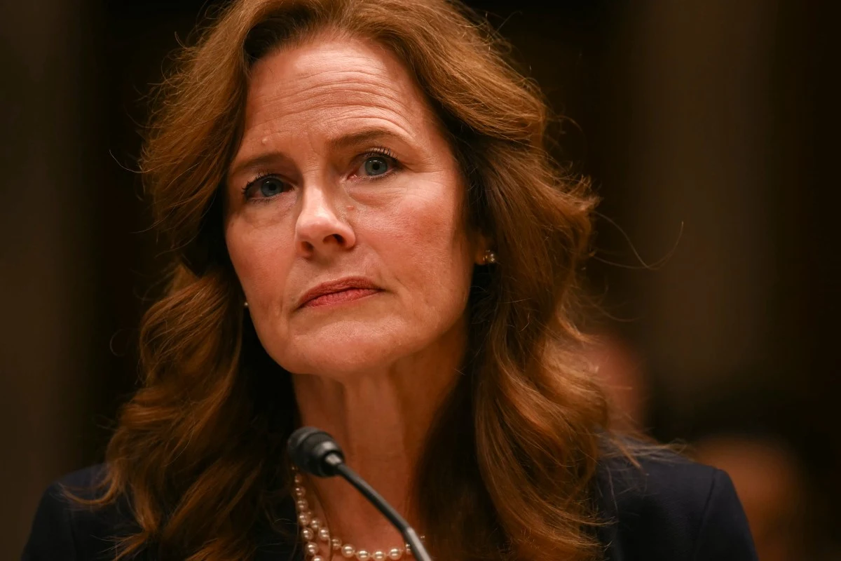 Amy Coney Barrett Kobieta o długich, falowanych włosach w eleganckim stroju siedzi za mikrofonem, ma poważny wyraz twarzy i nosi perłowy naszyjnik.