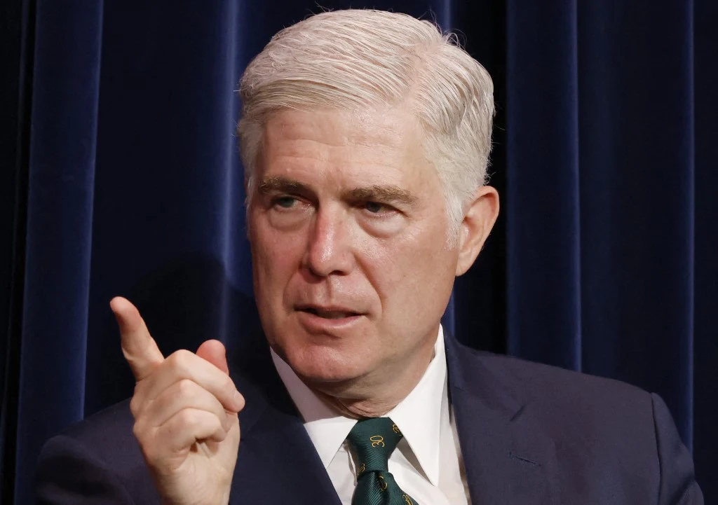 Neil Gorsuch Mężczyzna w garniturze z poważnym wyrazem twarzy, unosi dłoń z uniesionym palcem wskazującym, siedząc na tle ciemnych zasłon.
