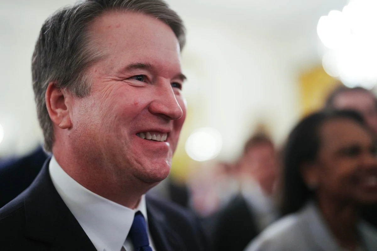 Brett Kavanaugh Uśmiechnięty mężczyzna w garniturze z krawatem, otoczony przez innych ludzi w eleganckich strojach, w jasnym wnętrzu.