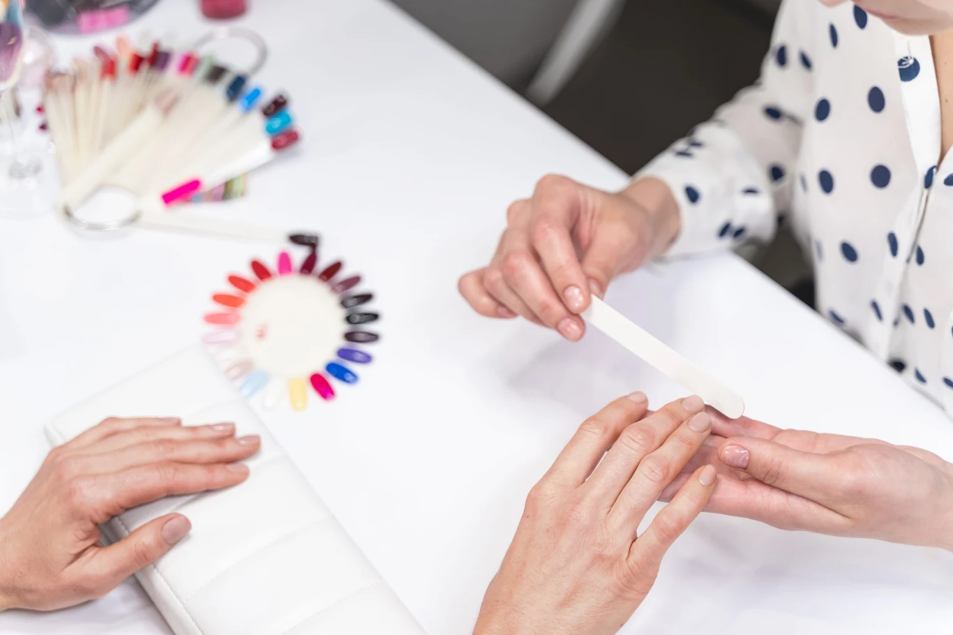 Na jaki wzór paznokci postawić jesienią? Ten jest teraz najmodniejszy Stylistka piłuje paznokcie klientki przy stole z próbnikiem kolorowych lakierów podczas manicure.